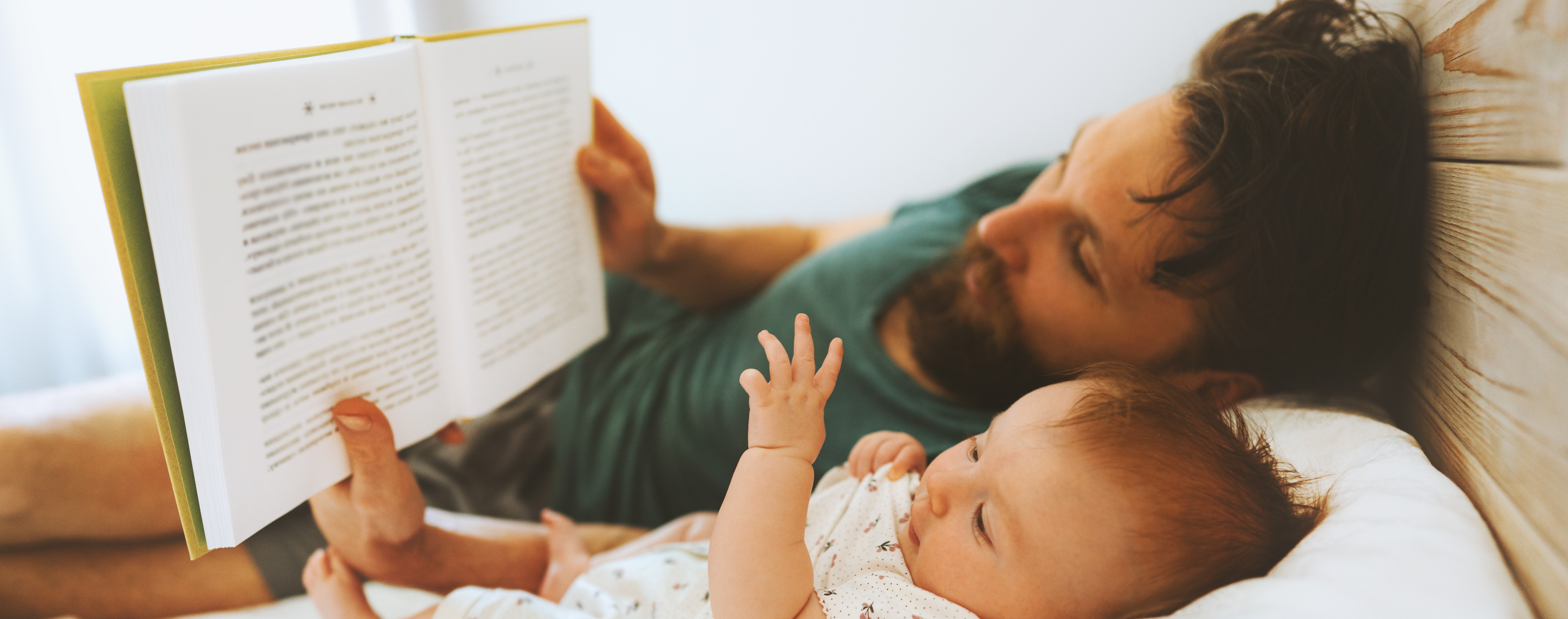 Picture of a father who is lying on a bed with a baby, appearing to read to it from a book.