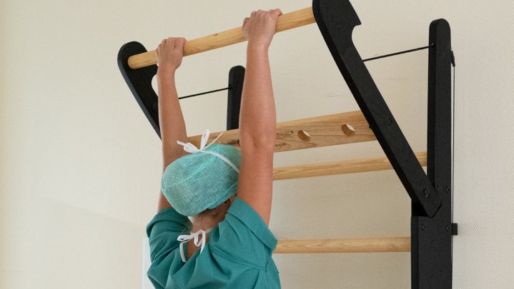 Eine Frau im grünen OP-Kasack und mit Schutzhaube hängt mit dem Gesicht zur Wand an der obersten Stange der Sprossenwand.