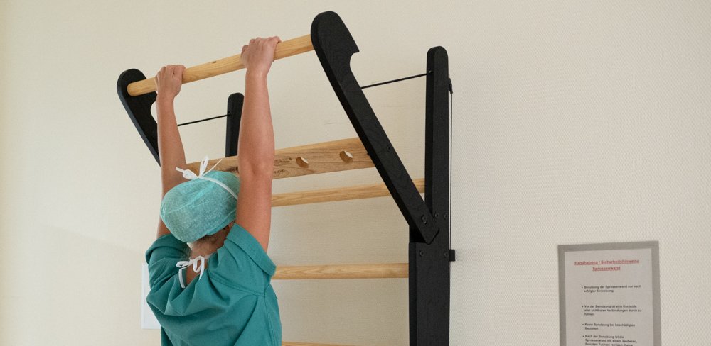 Eine Frau im grünen OP-Kasack und mit Schutzhaube hängt mit dem Gesicht zur Wand an der obersten Stange der Sprossenwand.