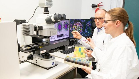 Ein Wissenschaftler und eine Wissenschaftlerin sitzen vor einem High-Tech-Mikroskop.