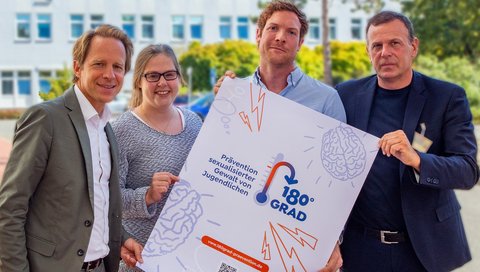 Professor Dr. Tillmann Krüger, Theresa Engelmann, Dr. Jonas Kneer und Klinikdirektor Professor Dr. Stefan Bleich schauen sich das Werbe-Poster zur neuen Ambulanz „180Grad“ an