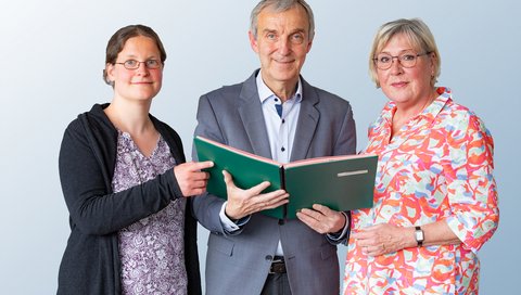 Ein Mann steht zwischen zwei Frauen und hält eine Aktenmappe in den Händen.