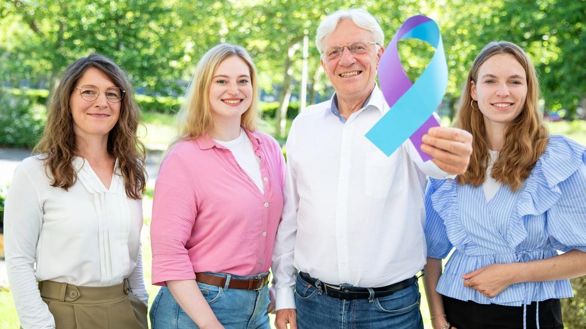 Dr. Kathrin Krüger, Viktoria Wiesner, Prof. Dr. Christian Krauth und Laura Böhme stehen für ein Gruppenbild zusammen im Freien. Prof. Krauth hält eine große blau-violette Schleife in Richtung Kamera.