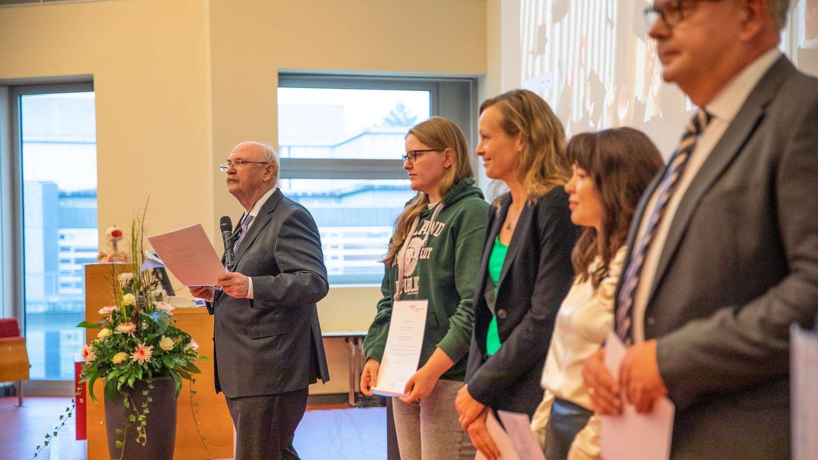 Professor Dr. Michael P. Manns steht mit Mikrofon auf dem Podium eines Hörsaals. Neben ihm stehen vier der ausgezeichneten Doktoranden und halten ihre Urkunden in den Händen. . 