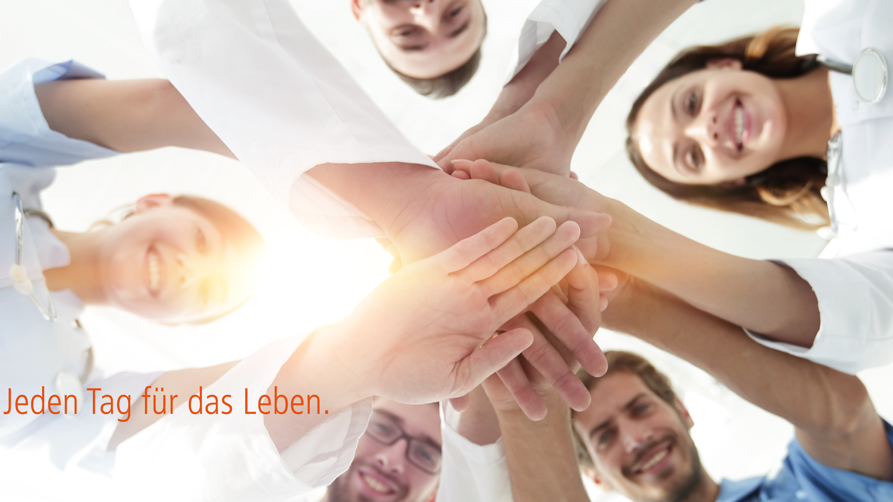 Mehrere Menschen stehen im Kreis und halten die Hände aufeinander und schauen nach unten in Richtung Kamera. 