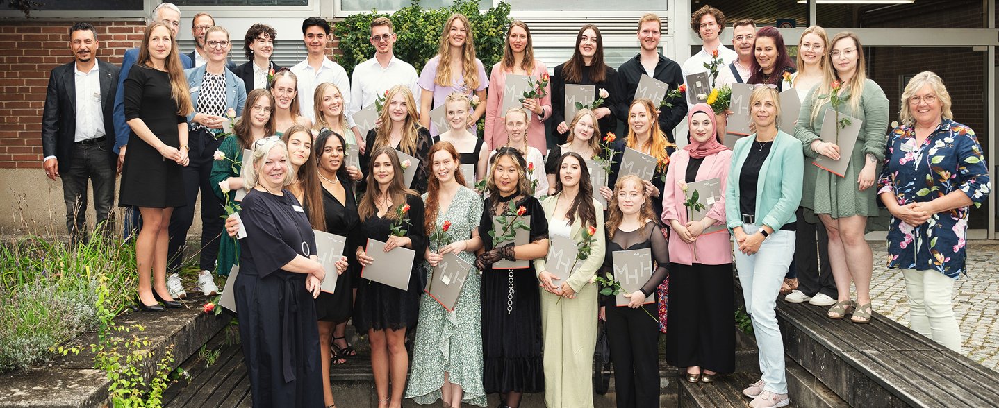 Mehrere junge Menschen stehen draußen zwischen Bänken vor der Bildungsakademie Pflege für ein Gruppenfoto in drei Reihen aufgestellt. Sie halten ihre Abschlussurkunden und Rosen in den Händen. 