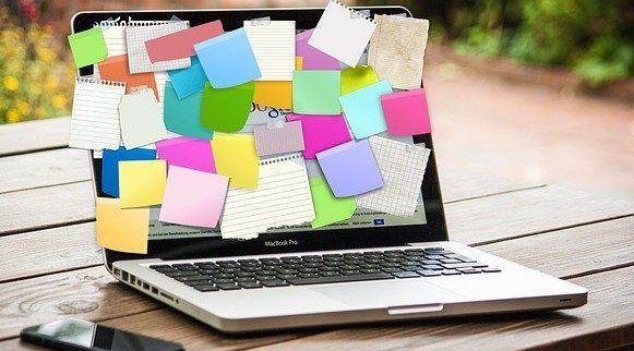 Copyright: Gerd Altmann/Pixabay_viele bunte Post-Its, die auf einem Laptop-Monitor aufgeklebt sind. Links neben dem Laptop liegt ein Mobiltelefon. Der Laptop steht auf einem Holztisch, im Hintergrund sind verschwommen Pflanzen zu sehen.