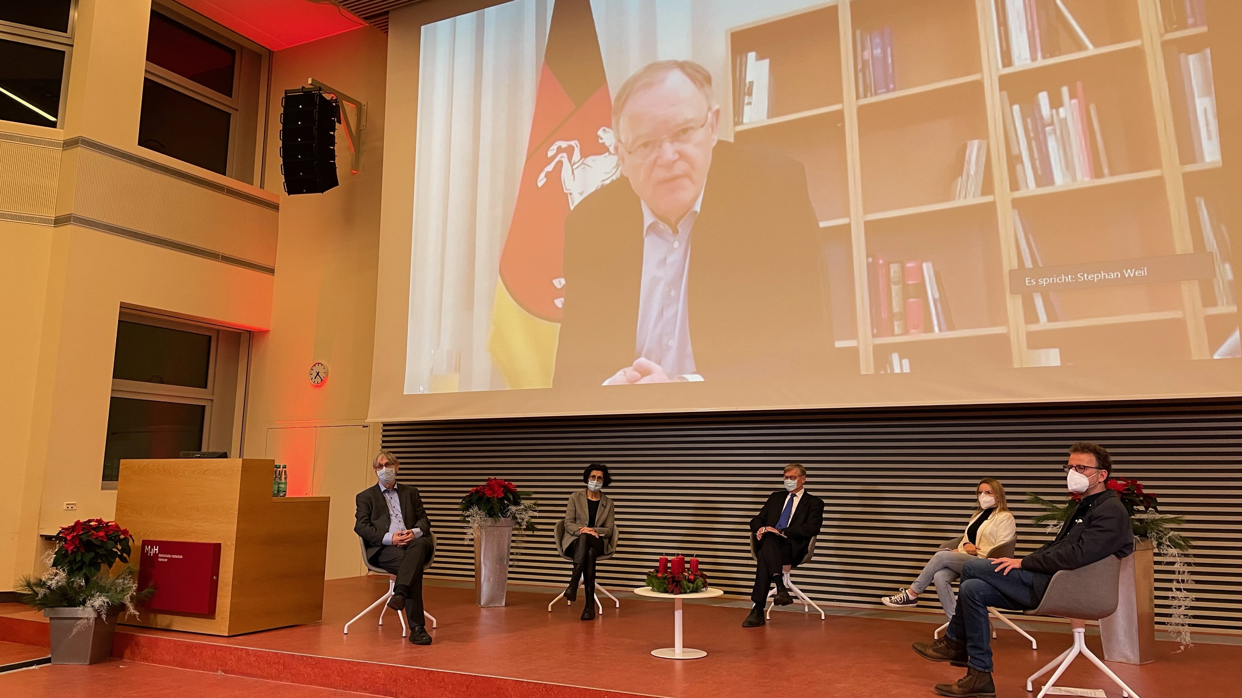 Auf dem Podium eines Hörsaals der MHH sitzen fünf Personen. Niedersachsens Ministerpräsident ist über eine Leinwand im Hintergrund dazugeschaltet. 