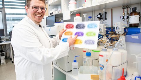 Ein Wissenschaftler im weißen Kittel steht in einem Forschungslabor und präsentiert eine bunte, symbolische Darstellung von Bakterien.