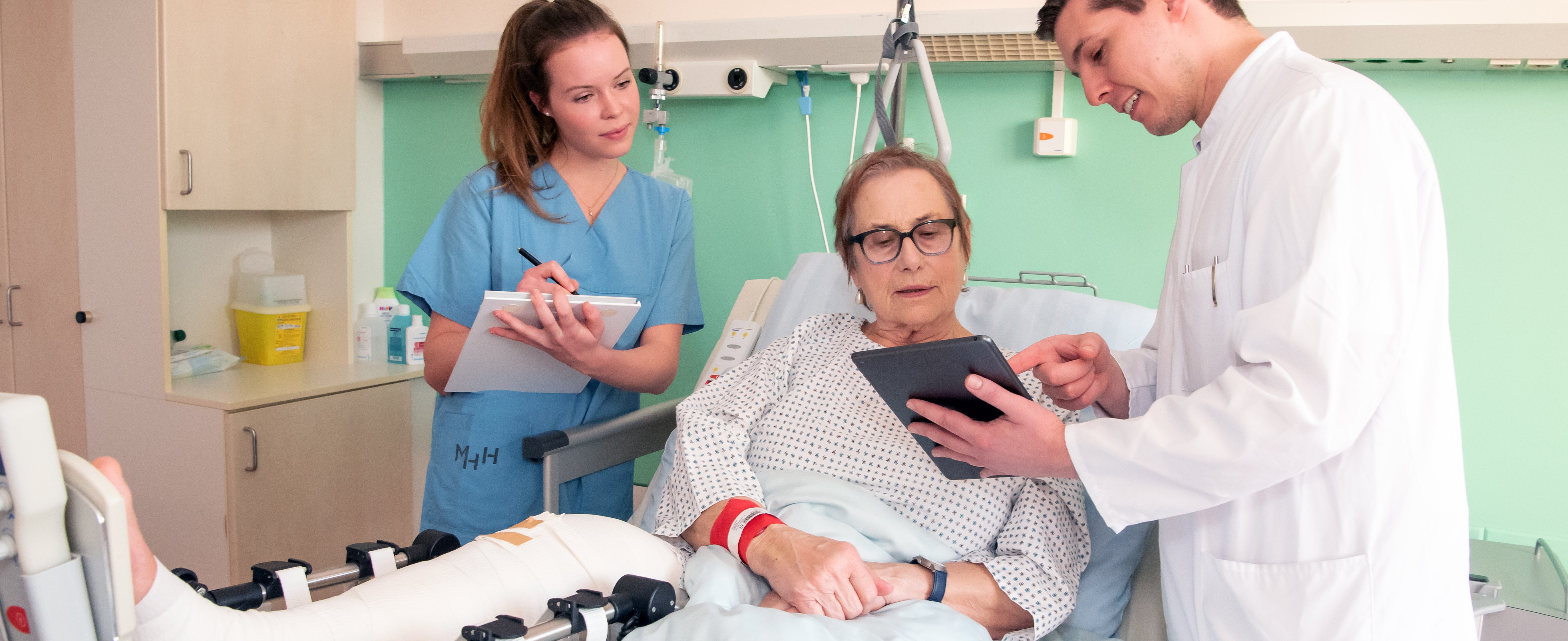 Eine Pflegerin und ein Arzt besprechen die Behandlung mit einer Patientin, deren Bein auf einer Bewegungsschiene liegt.