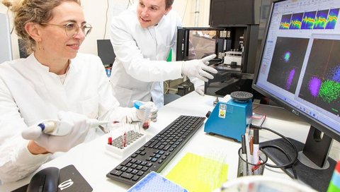 Ein Mann und eine Frau haben je einen Kittel an. Sie sind in einem Labor zu sehen an einem Bildschirm. 