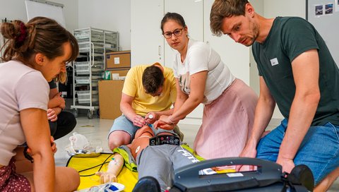 Studierende sitzen im Halbkreis an einer speziellen Übungspuppe, an der sie die Reanimation mit Hilfe eines Defibrillators üben. 