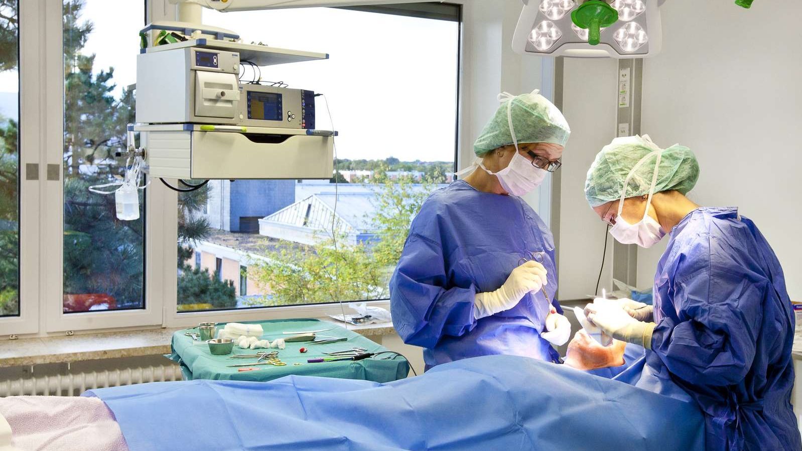 Zwei Ärzt:innen in OP-Kleidung stehen an einem OP-Tisch und beugen sich über einen dort liegenden Patienten. 
