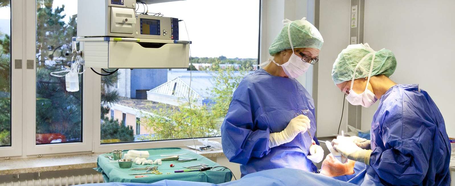 Zwei Ärzte in OP-Kleidung stehen am OP-Tisch und beugen sich über einen dort liegenden Patienten. 
