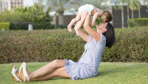A woman sits on a lawn and lifts a small child into the air.