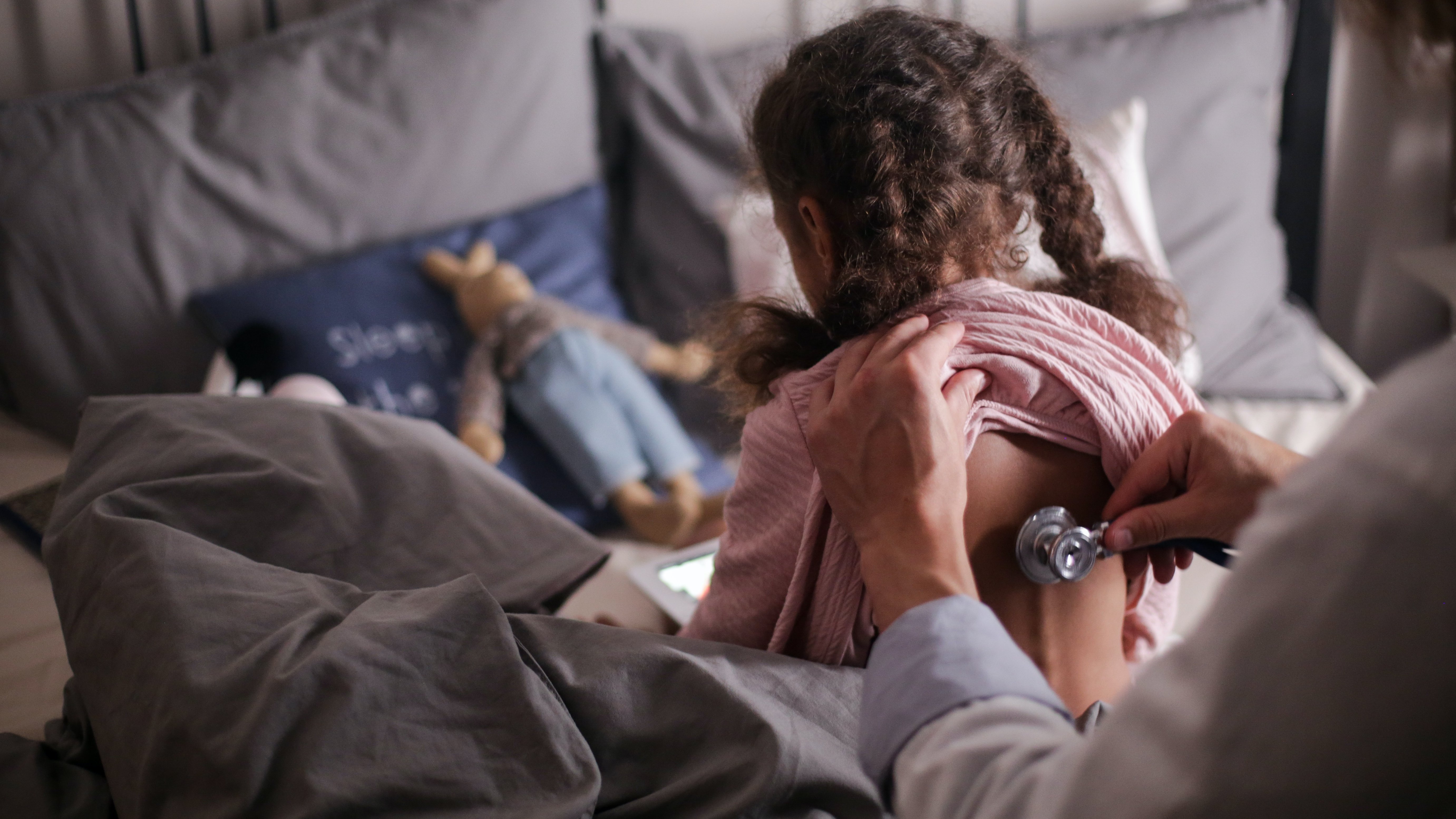 Ein kleines Mädchen sitzt auf einem Patientenbett und wird von einem Arzt mit einem Stethoskop am Rücken abgehört.