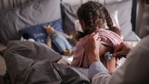 Ein kleines Mädchen sitzt auf einem Patientenbett und wird von einem Arzt mit einem Stethoskop am Rücken abgehört.