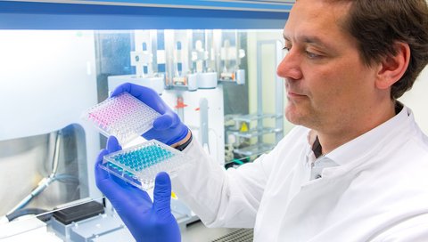 Professor Dr. Christian Bär stands in front of a pipetting robot and holds two micropipetting plates in his hands