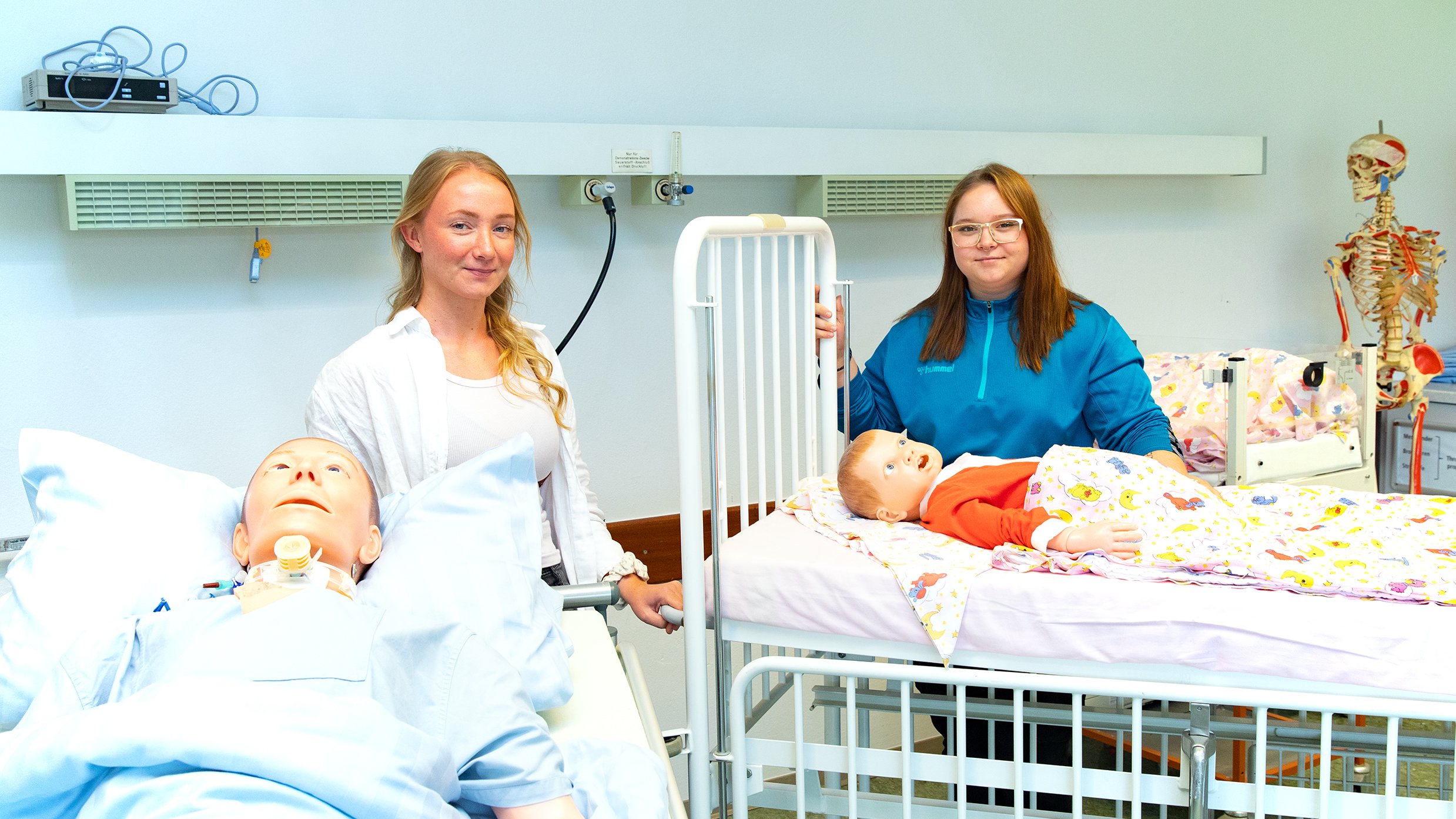 Marleen Schneider und  Rieke Busch-Quintero Dominguez stehen an zwei Betten mit  Puppen als Übungsdummys