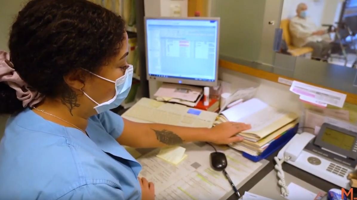 Medizinische Fachangestellte sitzt mit Mundschutz am Schreibtisch. 
