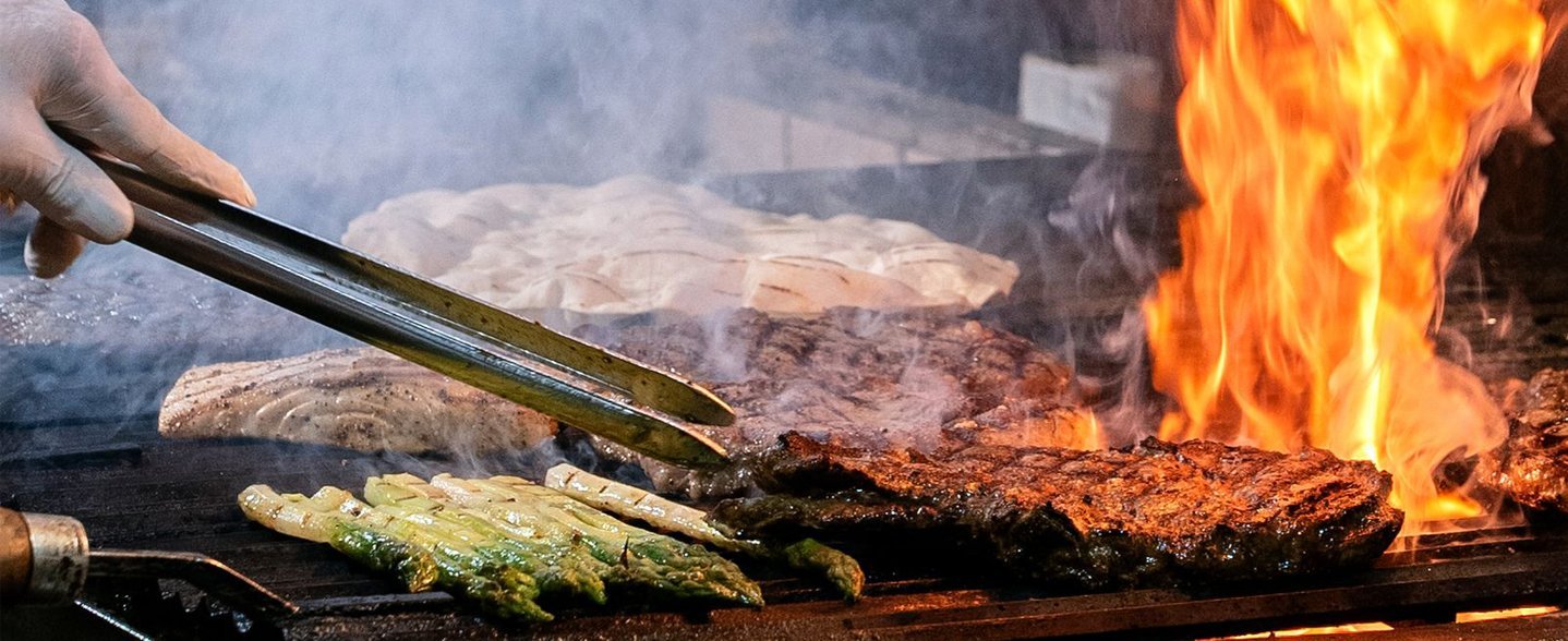Nahaufnahme eines Grilles mit Fleischstücken und einer lodernden Flamme. Eine Hand hält eine Grillzange.  