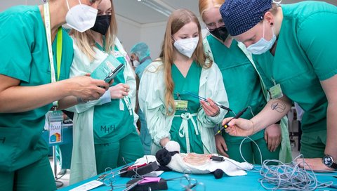 Mehrere junge Menschen in grüner OP-Schutzkleidung stehen um eine Tisch, auf dem ein Schweineschnitzel liegt. Eine Person hält einen Metallstab in der Hand. 