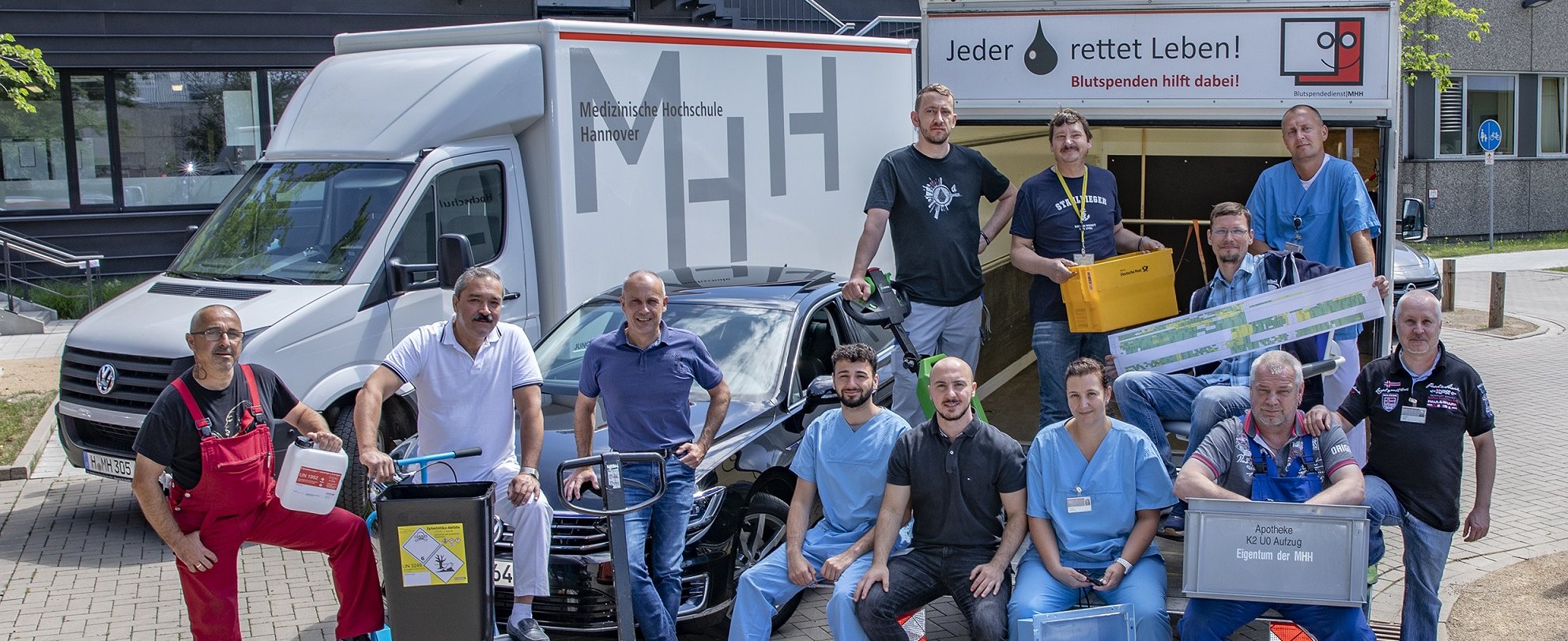 Gruppenbild von Mitarbeitern der Abteilung Transportwesen und Materialwirtschaft mit verschiedenen Arbeitsmaterialien und im Hintergrund drei Fahrzeuge der MHH