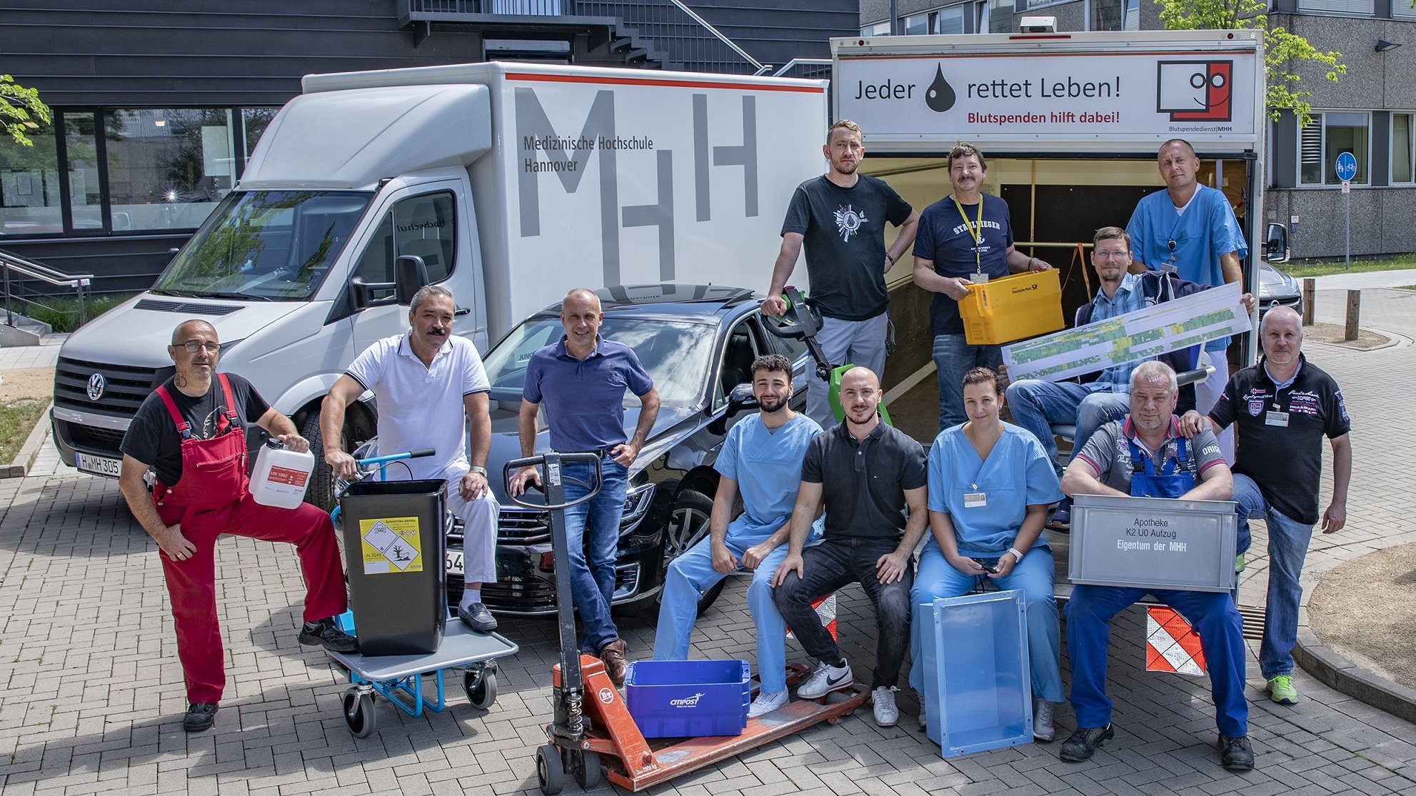 Gruppenbild von Mitarbeitern der Abteilung Transportwesen und Materialwirtschaft mit verschiedenen Arbeitsmaterialien und im Hintergrund drei Fahrzeuge der MHH