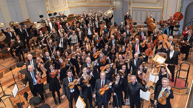 Die Musikerinnen und Musiker der Bundesärztephilharmonie stehen in einem prunkvollen Konzertsaal und halten ihre Instrumente in die Höhe.