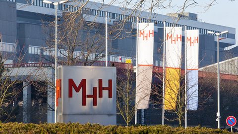 Die Metallstele an der Haupteinfahrt mit den roten Buchstaben MHH. Dahinter ein Gebäude mit drei Fahnen mit MHH-Aufschriften. 