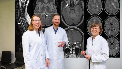 Franziska Bütow, Dr. Hümmert und Professorin Trebst, alle drei in weißem Kittel, stehen vor einer großen Aufnahme des Gehirns.