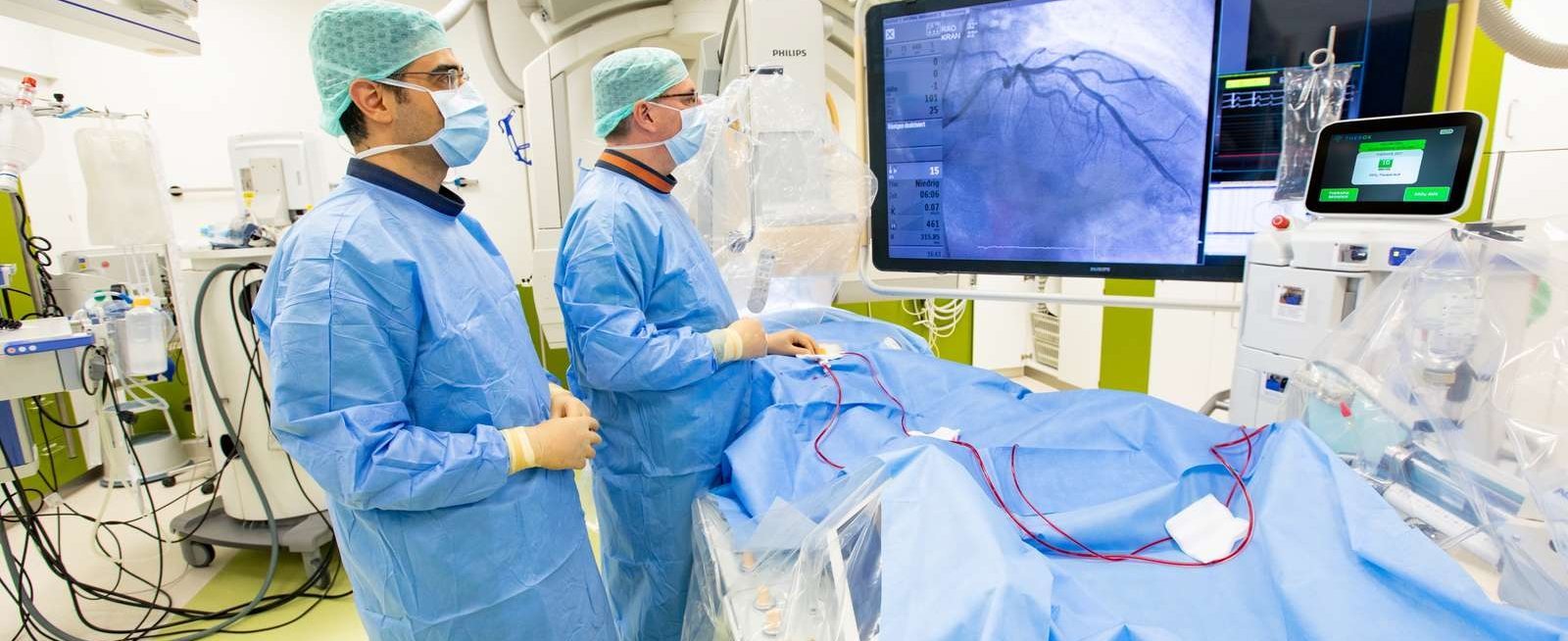 Zwei Männer in OP-Schutzkleidung stehen in einem OP am Operationstisch, auf dem ein Patient mit blauem Laken zugedeckt liegt, und betrachten den Monitor über dem OP-Tisch. 