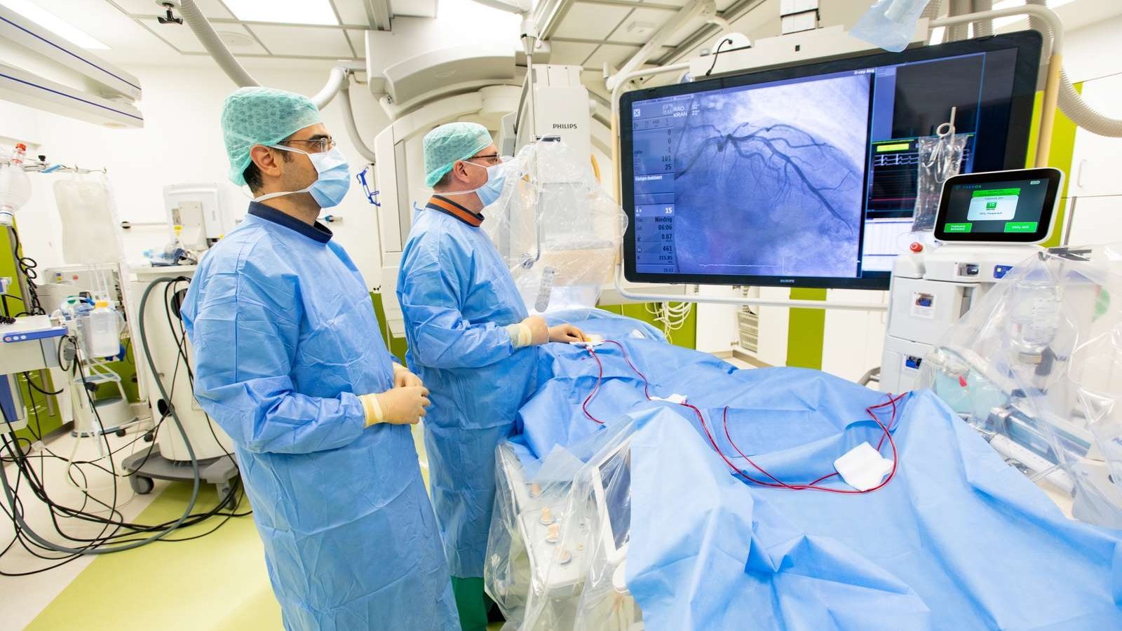 Zwei Männer in OP-Schutzkleidung stehen in einem OP am Operationstisch, auf dem ein Patient mit blauem Laken zugedeckt liegt, und betrachten den Monitor über dem OP-Tisch. 
