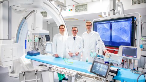 Drei Männer stehen an einer Herzkatheteranlage.