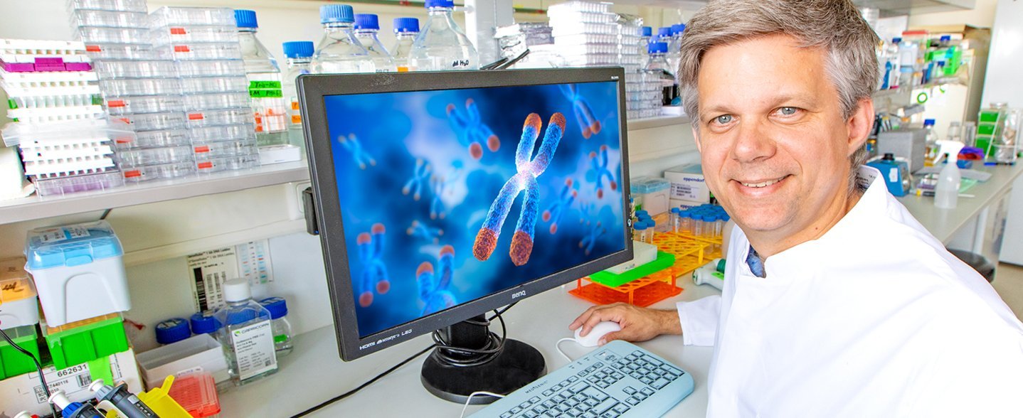 Auf dem Foto ist Prof. Dölken im weißen Laborkittel zu sehen, er sitzt auf einem Stuhl in einem Labor und auf dem Tisch neben ihm steht ein Bildschirm.