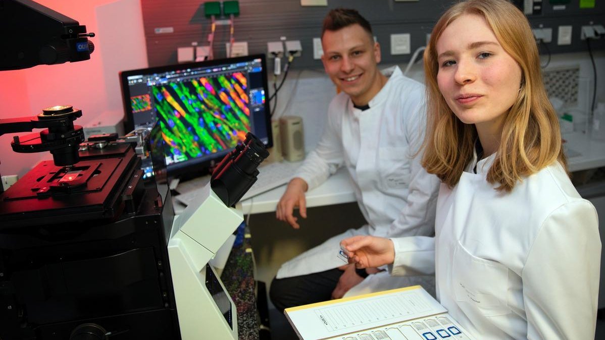 Ein Mann und eine Frau in weißen Laborkitteln sitzen vor einem Monitor. Beide blicken in die Kamera.