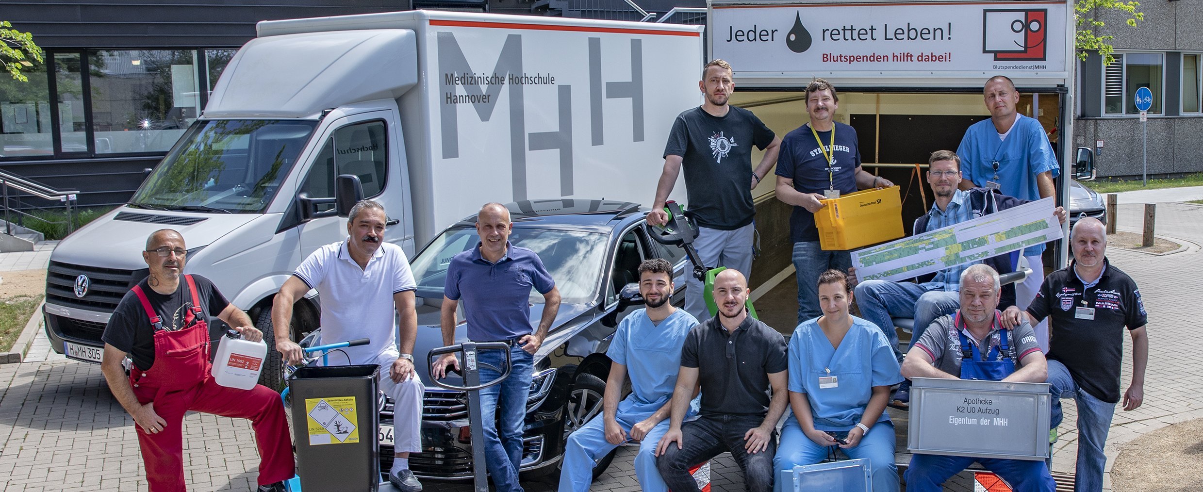 Gruppenbild von Mitarbeitern der Abteilung Transportwesen und Materialwirtschaft mit verschiedenen Arbeitsmaterialien und im Hintergrund drei Fahrzeuge der MHH