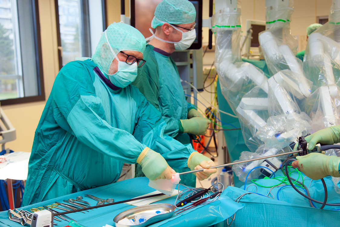 Op-Pflegekraft im Vordergrund, dahinter Chirurg, beide steril eingekleidet, im Hintergrund 3 der 4 Arme des OP-Roboters