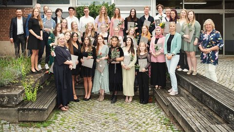 Mehrere junge Menschen stehen draußen zwischen Bänken vor der Bildungsakademie Pflege für ein Gruppenfoto in drei Reihen aufgestellt. Sie halten ihre Abschlussurkunden und Rosen in den Händen.  