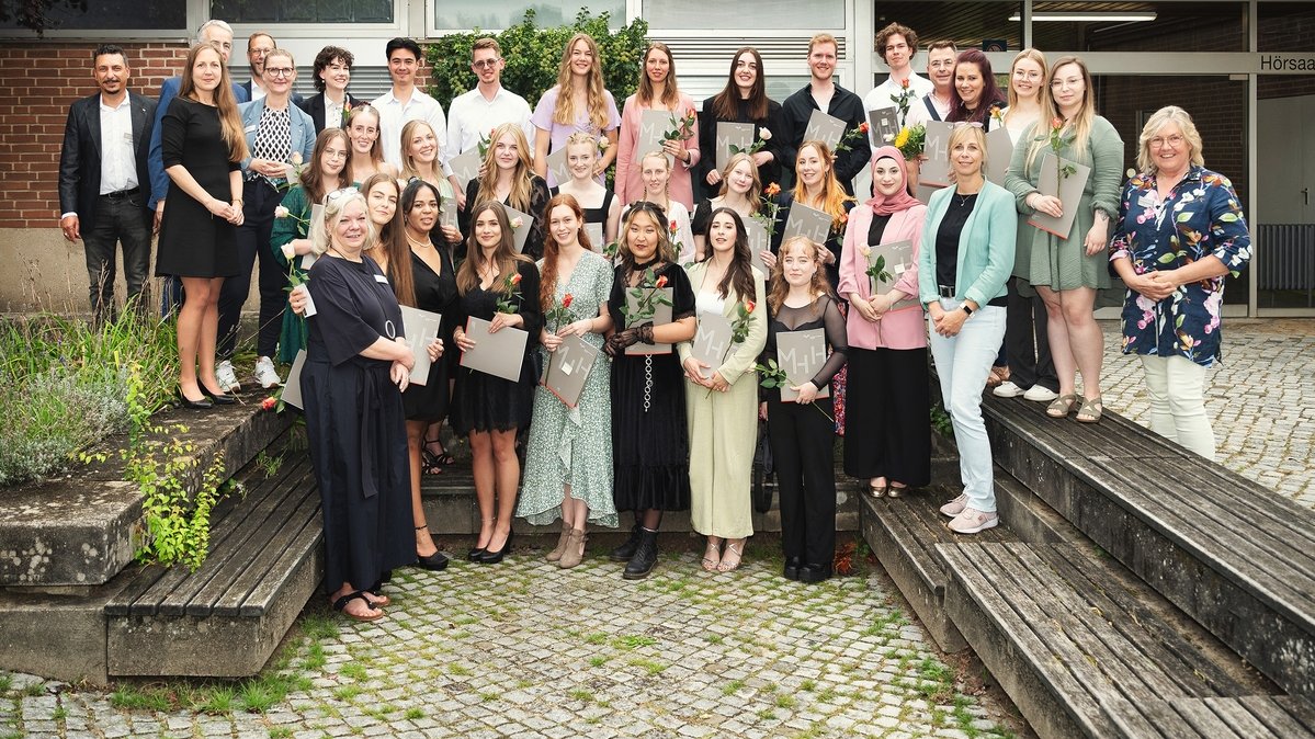 Mehrere junge Menschen stehen draußen zwischen Bänken vor der Bildungsakademie Pflege für ein Gruppenfoto in drei Reihen aufgestellt. Sie halten ihre Abschlussurkunden und Rosen in den Händen.  