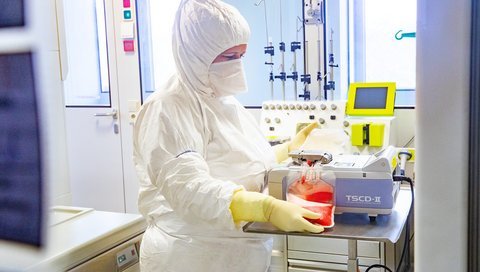 A person in full protective clothing stands at a table in a laboratory and operates a device with a filled blood plasma bag.