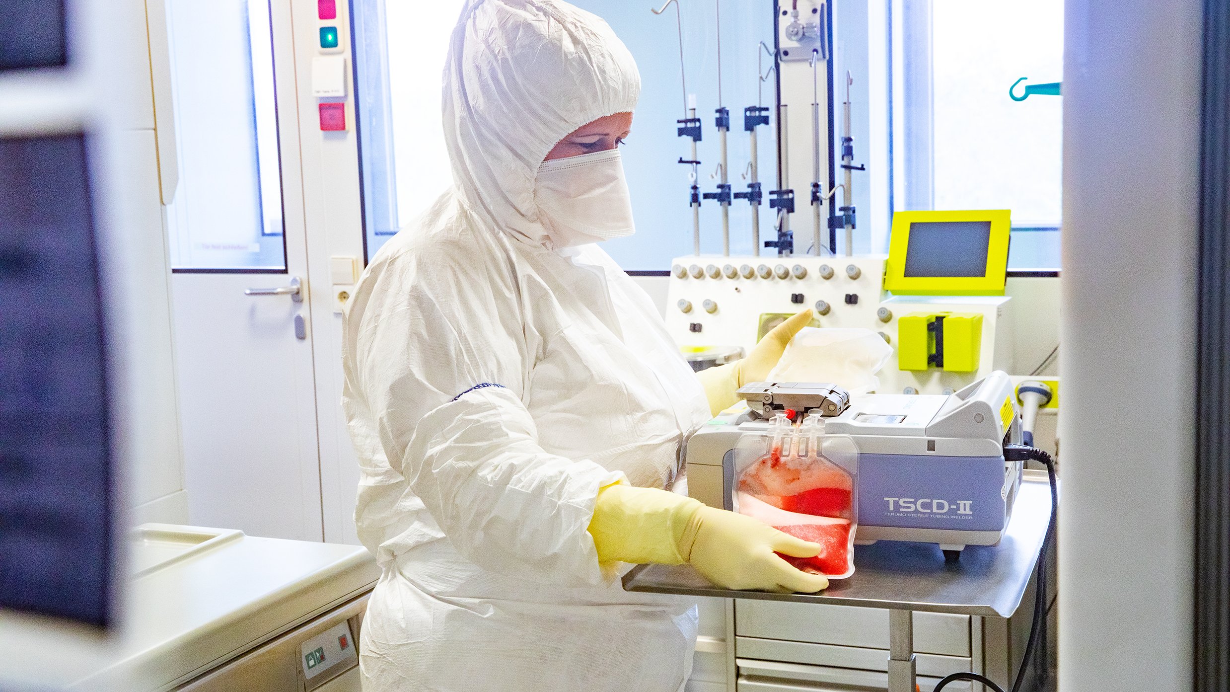 A person in full protective clothing stands at a table in a laboratory and operates a device with a filled blood plasma bag.