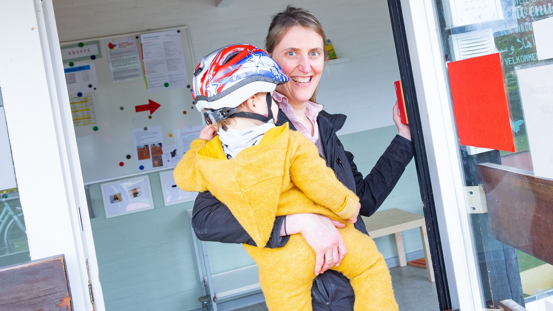 Eine Frau trägt ihr Kind auf dem Arm. Das Kind trägt eine gelben Anzug und einen Fahrradhelm.