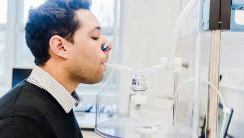 Mann sitzt vor Inhalationsgerät mit Mundstück im Mund und zugeklammerter Nase. 