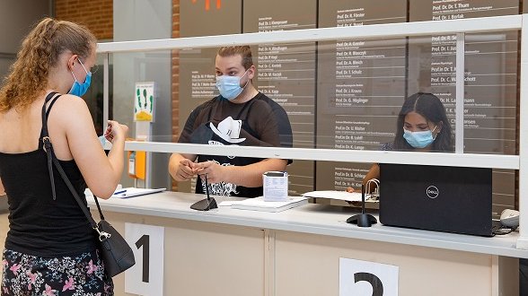 Eine Frau mit Mundschutz steht an einem Empfangstresen mit Glasscheibe, hinter dem ein Mann und eine Frau ebenfalls mit Mundschutz stehen. 