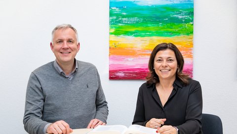 Professor Szycik und Professorin Zimmermann sitzen an einem Tisch und unterhalten sich über den neuen Masterstudiengang, im Hintergrund ist ein buntes Bild an der Wand.