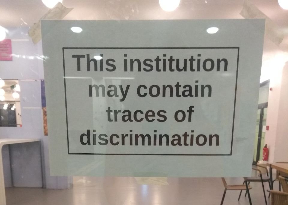 Durch eine Glasscheibe fotografiertes Papier mit der Aufschrift "This institution may contain traces of discrimination"
