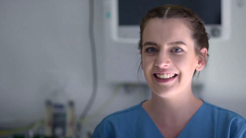 Porträtbild von Kinderkrankenpflegerin Pia, die einen blauen Kasack trägt und in einem Patientenzimmer mit Monitoren im Hintergrund steht. 