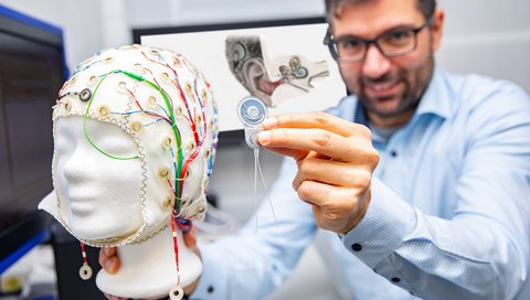 Professor Dr. Waldo Nogueira Vazquez hält ein Cochlea-Implantat an eine EEG-Haube zur Messung der Signale bei der Hörverarbeitung im Gehirn