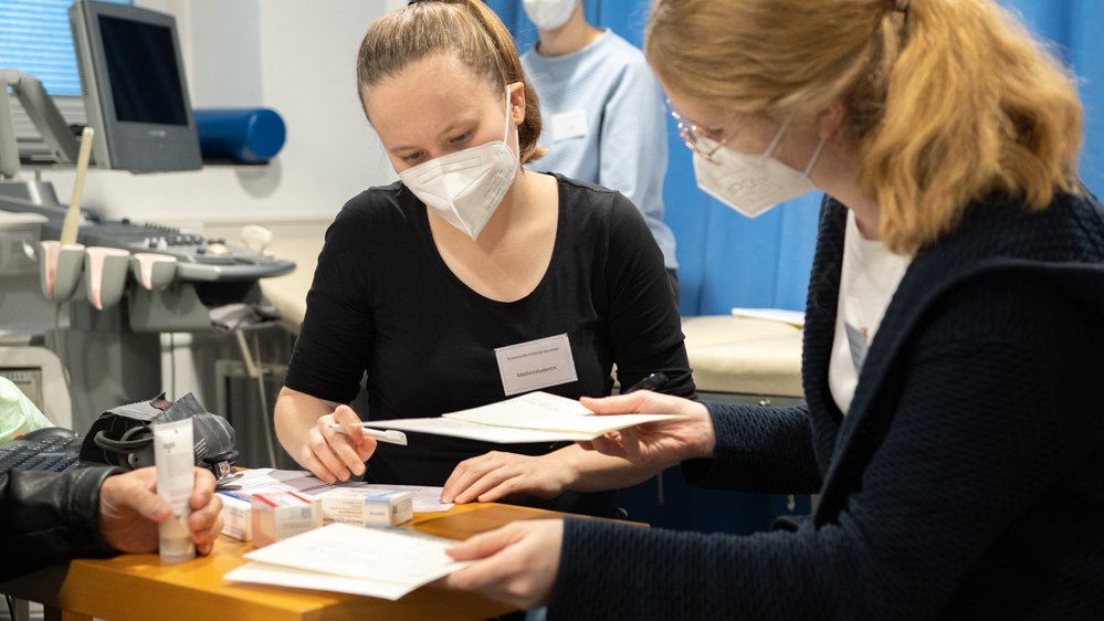 Zwei Studentinnen beraten im Vordergrund einen Patienten, im Hintergrund sitzt eine Ärztin auf einer Behandlungsliege.